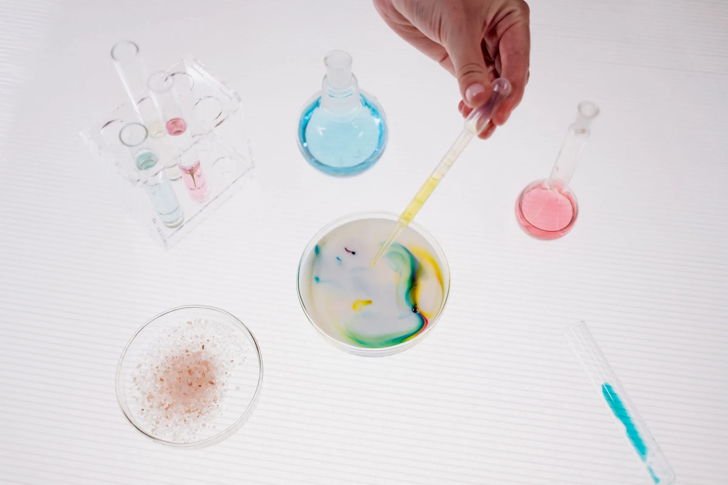A hand dropping colour from a pipette into a dish, among flasks of tinted liquid